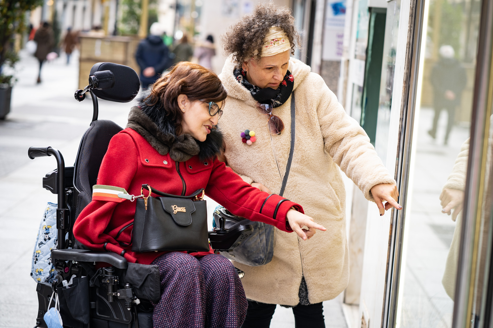 Una mujer en silla de ruedas con su asistente personal en la calle mirando escaparates.
